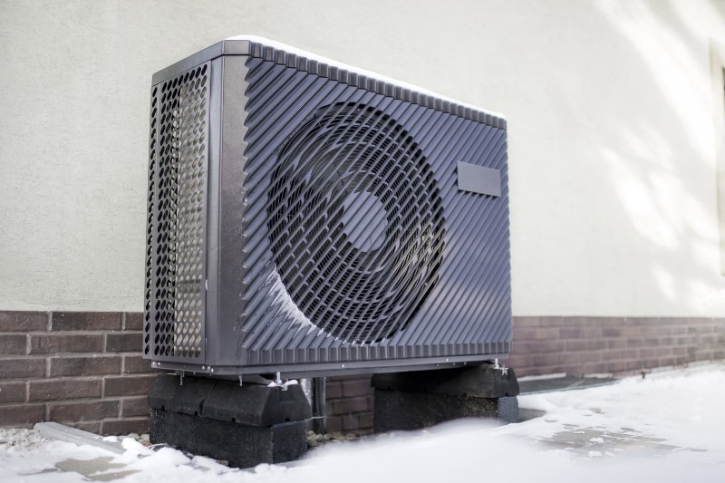 How to Determine Heat Pump Efficiency. Photo of a modern air source heat pump unit standing against the residential building wall. Shot in winter, in a snowy setting.