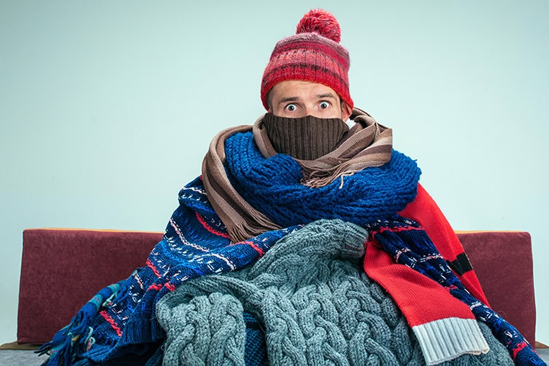 How Can I Make My Furnace Last Longer? A woman sitting on a couch bundled up in blankets and scarves.